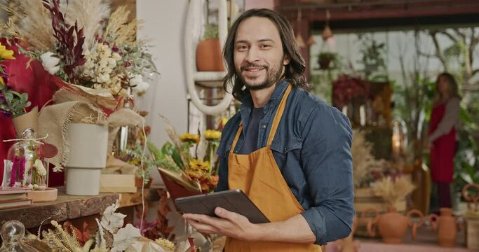 Portrait of a male worker wearing apron and holding tablet turns head towards camera smiling inside retail store shop. Small business concept