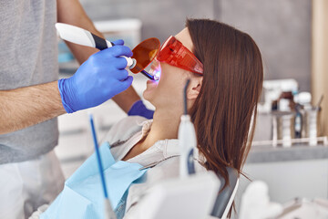 Professional male doctor uses ultraviolet lamp after making a filling on a tooth for joyful happy caucasian woman in modern dental cabinet