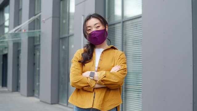 Portrait of young chinese woman in medical face mask standing near moden grey building with crossed arms and looking at camera. People, virus and prevention concept.