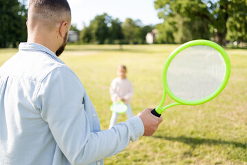 Rear view of man holding badminton racket while playing with his daughter outdoors in the park on a summer day