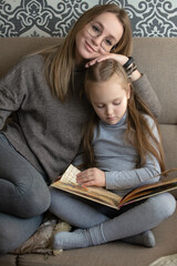 A little girl is reading a book at home on the couch, her mother next to her is hugging her