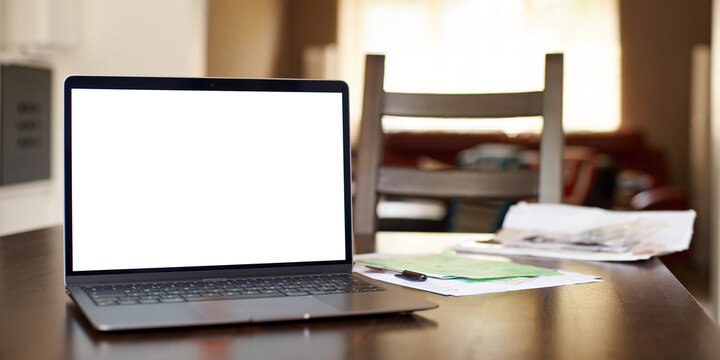 Mockup image of laptop with blank white desktop screen on a wooden table in an ordinary apartment. Business papers lie near the laptop in the home office. Laptop with blank screen for design.