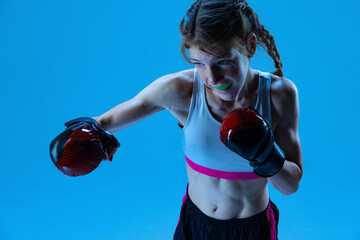 Dynamic portrait of young girl, junior MMA fighter in action, motion isolated on blue background in...