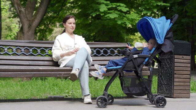 Mother with little cute child in baby stroller in the park, woman is resting on a bench on a sunny day, baby is drinking milk formula from a baby bottle.