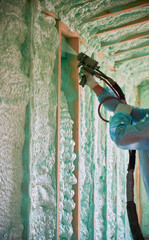 Builder insulating wooden frame house. Close up view of man worker spraying polyurethane foam inside of future cottage, using plural component gun. Construction and insulation concept.