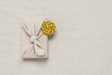 Gift wrapped in fabric with lollipop, beige linen textile background. Furoshiki gift. Zero waste concept. Reusable sustainable gift wrapping. Top view, flat lay.