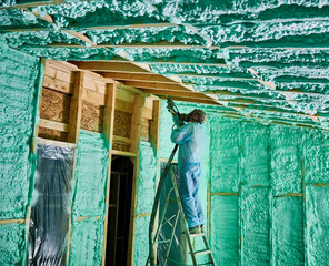 Male builder insulating wooden frame house. Man worker spraying polyurethane foam inside of future...