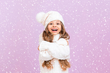 A little girl in a knitted white sweater and hat hugs herself and tries to keep warm smiling broadly . A child on a pink background in a snowstorm.