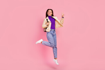 Full length photo of nice brunette lady jump show v-sign wear jacket jeans shoes bag isolated on pink color background