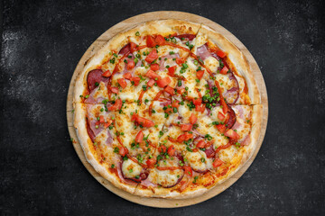 Pizza with salami ham tomatoes and hot spicy sauce, on a wooden round board on gray concrete. top view