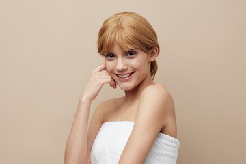 a sweet, elegant woman stands on a beige background wrapped in a white towel and smiling pleasantly at the camera holds her hand near her face