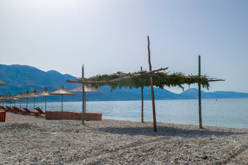 Naklejka na ściany i meble Beach umbrellas, and sun loungers on pebble beach. Qeparo village. Albania. Ionian Sea. Vacation sea concept. Summer sunny seaside landscape.
