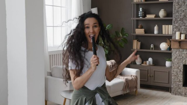 Active funny young woman looking at camera excitedly singing song holding tv remote as microphone having fun at home on weekends amuses dancing vigorously energetic moving to favorite incendiary music