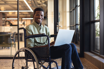 Smiling african american businessman with disability using laptop in wheelchair at workplace - obrazy, fototapety, plakaty