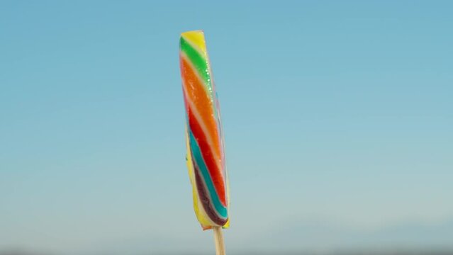 Multicolored lollipop on a stick, rotating against a blue sky background, close-up.