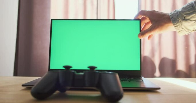 Hand closes the lid of laptop with green screen. Desktop, gamepad controller lies on tabletop. Concept of the end of the game, work, while sleeping, take a break from the computer