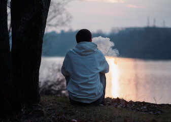 A young man in a white hoodie sits on the riverbank at sunset and exhales smoke from an electronic cigarette. Vaping man