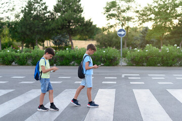 Two children look at their mobile phones while crossing a crosswalk on the street. Concept of mobile phone addiction - obrazy, fototapety, plakaty