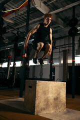 Portrait of sportive, muscular red-haired young man jumping on box, doing exercises isolated over gym background