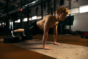 Fototapeta na wymiar Portrait of young red-haired sportive man with muscular body training, doing push-ups exercises isolated over geym background