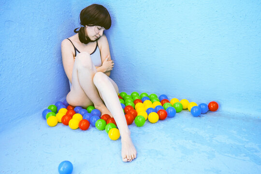 Sad woman sitting amidst multi colored balls by blue swimming pool wall