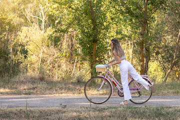 Fototapeta premium Side view of young elegant woman getting on retro pink bike in the middle of nature in spring time