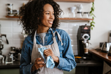 Black barista woman wearing apron wiping glasses while working in cafe