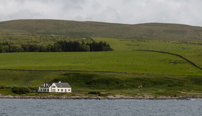 Isolated white house on Scottish shoreline with rolling fields of pasture land behind it.