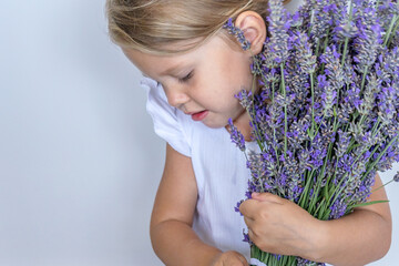 Shy white caucasian girl 2 years old with blond hair with a bouquet of lavender in her hands in a white dress on a white background during the holiday. Copy space for your text