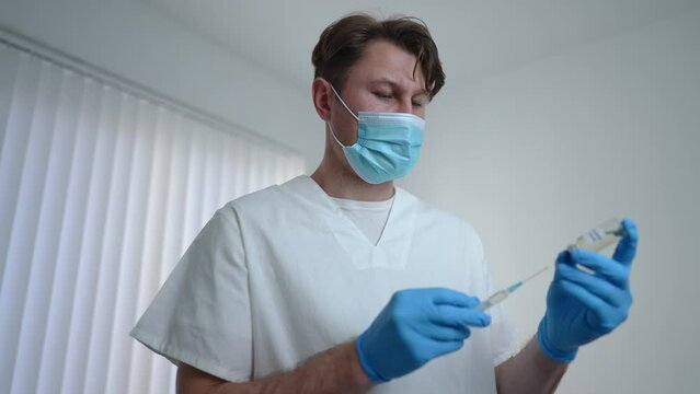 Concentrated serious doctor filling syringe with coronavirus vaccine standing in vaccination center. Portrait of confident focused Caucasian man in uniform and face mask preparing shot in hospital