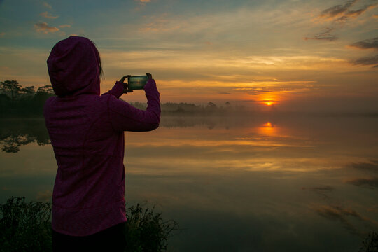Young woman traveller taking a picture of sunrise or sunset at the lake. She using the smartphone to capture a good moment of her vacation.