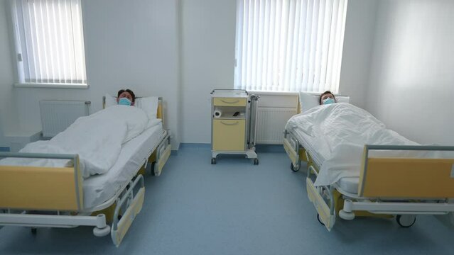 Wide shot two ill patients in coronavirus face masks lying in beds in hospital ward coughing. Unwell Caucasian young sick men in medical clinic on Covid-19 pandemic outbreak. Health and treatment