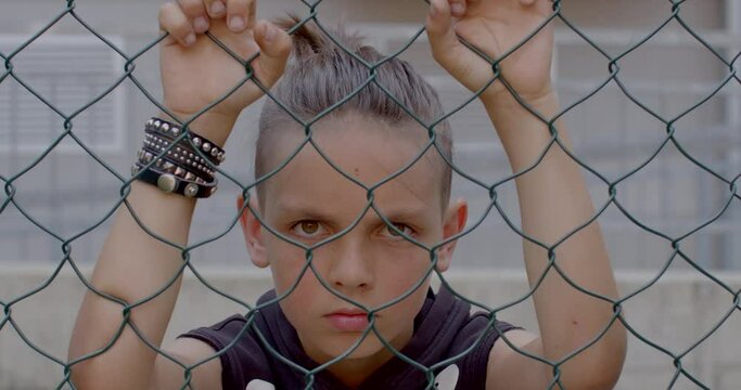 Angry child keeps his hands on the wall of the fence. He looks through the wire cages of the fenced area. Waiting for release. Rivet leather strap on hand.
