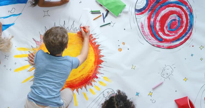 Young children and friends paint colorful space pictures indoors on the floor in kindergarten from the above view. Kids being creative painting science fiction drawings with colors on a big sheet.