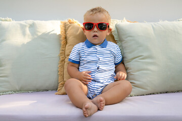 little boy 1 year old wearing sunglasses sits on a sofa outdoors.