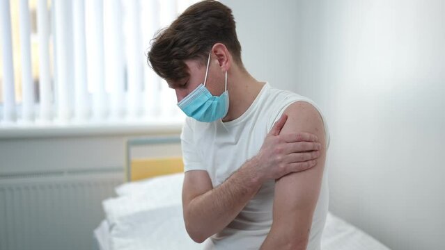 Side view portrait of sad Caucasian man in Covid-19 face mask with medical patch on shoulder sitting on bed in hospital ward. Unwell patient with severe ache in arm after coronavirus vaccination