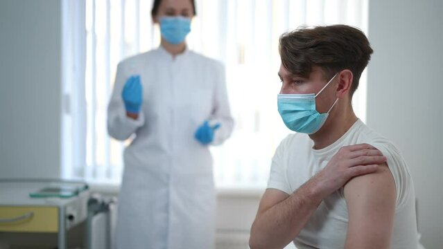 Scared Adult Man In Covid-19 Face Mask Looking At Camera Shacking Head No As Doctor Walking From Background With Coronavirus Vaccine Shot In Syringe. Vaccination And Fears Concept