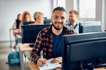 Happy student writing notes while using desktop PC on computer class and looking at camera.