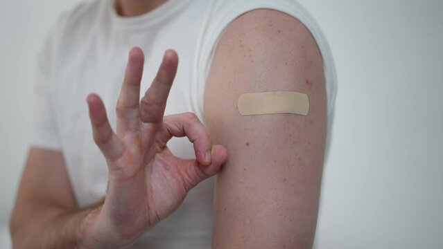 Close-up male hand gesturing OK at shoulder with medical patch after Covid-19 vaccination. Unrecognizable adult Caucasian man getting vaccine shot in hospital. Illness prevention concept