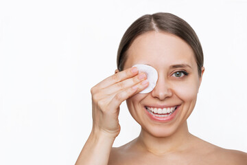 Close up of a young caucasian smiling woman cleaning her face with cotton pad removing makeup from the eyes isolated on a white background. Skin care, cosmetology. The girl washes off her makeup