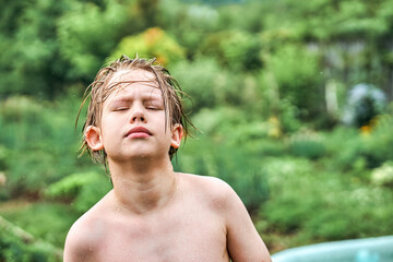 Cute schooler with wet blond hair stands against blurry lush trees and greenery in countryside....