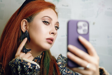 Cute girl takes selfie on front camera of modern smartphone. Female blogger films herself for fashion and makeup blog. Red-haired woman points her finger at her butterfly earrings. Gothic aesthetic.