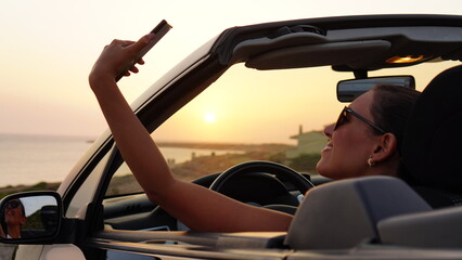 Attractive woman makes selfie and takes photos of the sunset from her convertible car parked in front of the beach, enjoying the sunset alone and relaxing with the view