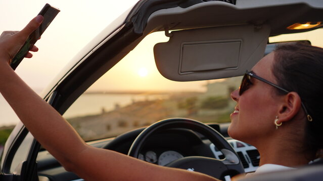 Attractive woman makes selfie and takes photos of the sunset from her convertible car parked in front of the beach, enjoying the sunset alone and relaxing with the view