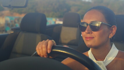 Woman alone in a black convertible car looks at the sunset in front of the sea and is moved by how...