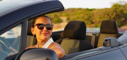 Woman alone in a black convertible car looks at the sunset in front of the sea and is moved by how wonderful it is, she feels free and happy during her vacation alone, independent and strong woman