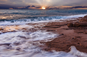 Obraz premium Beautiful colorful sunset over the sea with waves and rocky coastline. Peaceful Landscape with setting sun over the horizon