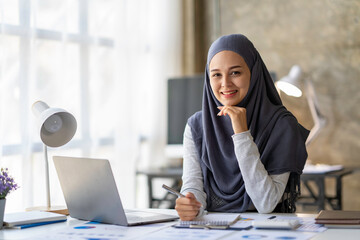 Attractive Muslim woman wearing hijab looks at camera working in business office. Use a calculator and laptop to calculate money on the desk, in the office, taxes, accounting, financial concepts.