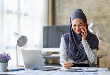 Attractive Muslim businesswoman wearing hijab Muslim in smart casual wear talking by phone while working in office discussing business and smiling while sitting and using the laptop for work online.