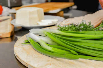 On the table are cheese, bread in a basket, herbs, dill, green onions, a knife on the board